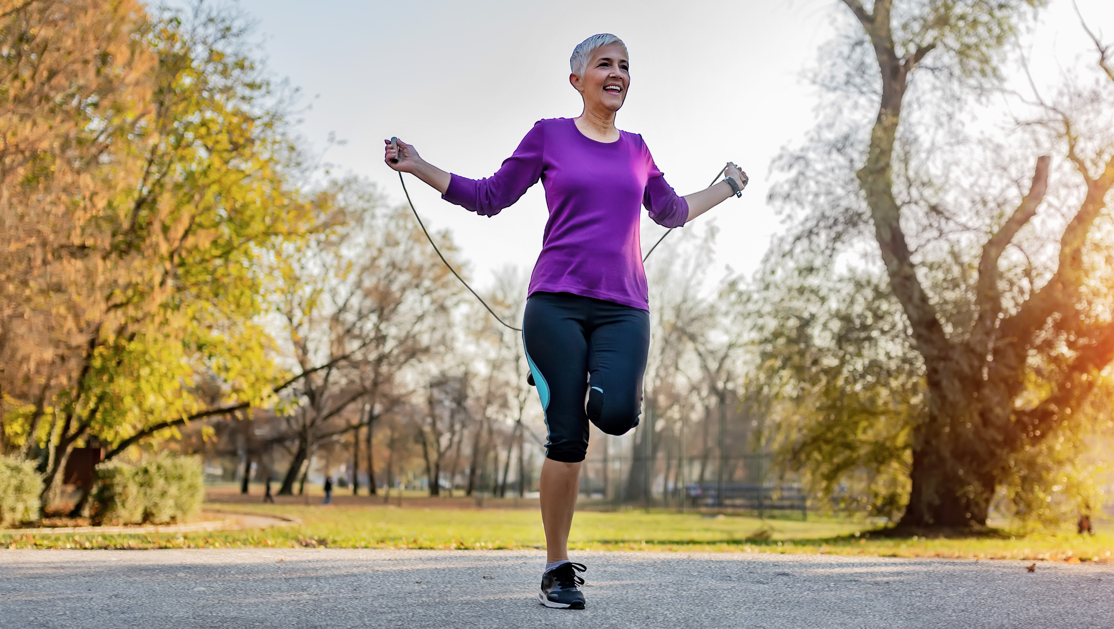 A woman jumps rope in a park. She is smiling and behind her we see trees and grass.