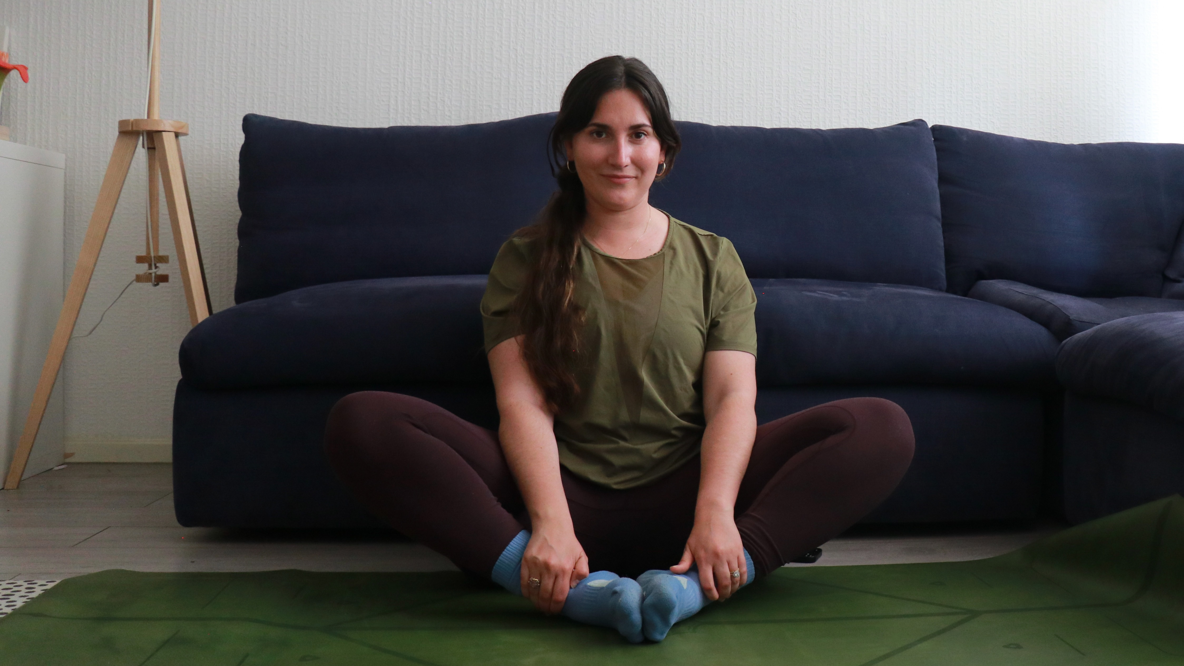 Marina Leiva Garc&iacute;a sits on a yoga mat performing a butterfly pose. She is sitting in front of a couch, with her knees bent and out to the sides and the soles of her feet touching. She holds her ankles in her hands.