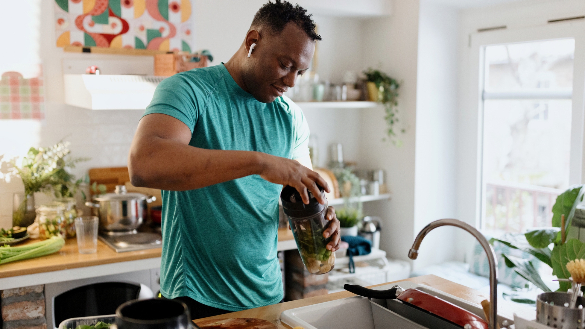 man in a turquoise tshirt in a kitchen by the sink holding a blender cup screwing the lid on. 