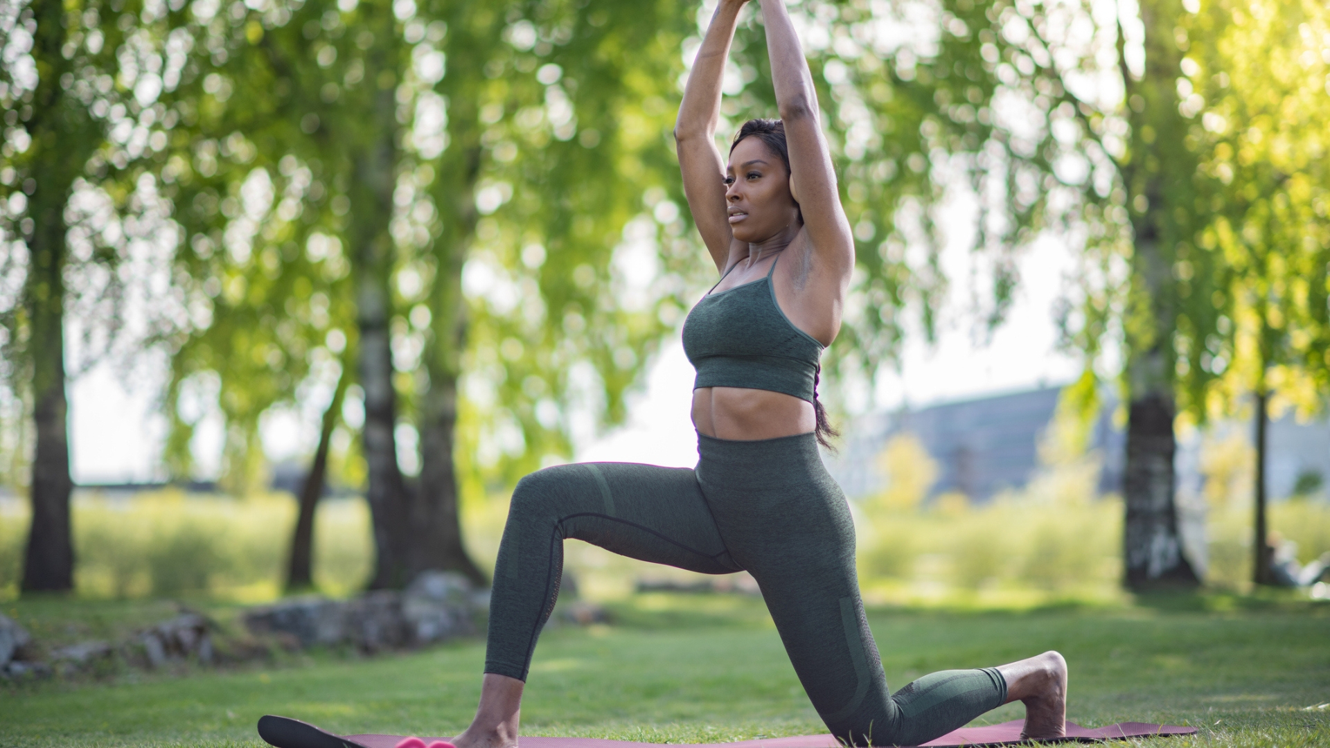 woman wearing khaki crop top and leggings performing a deep lunge with arms stretched overhead. she's in a park setting with trees behind her.
