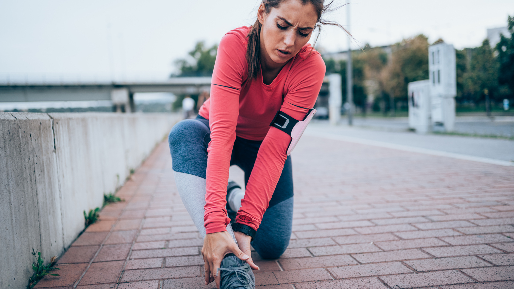 Woman dressed in long-sleeve running top and grey leggings kneels on one knee and holds her ankle with a look of concern on her face