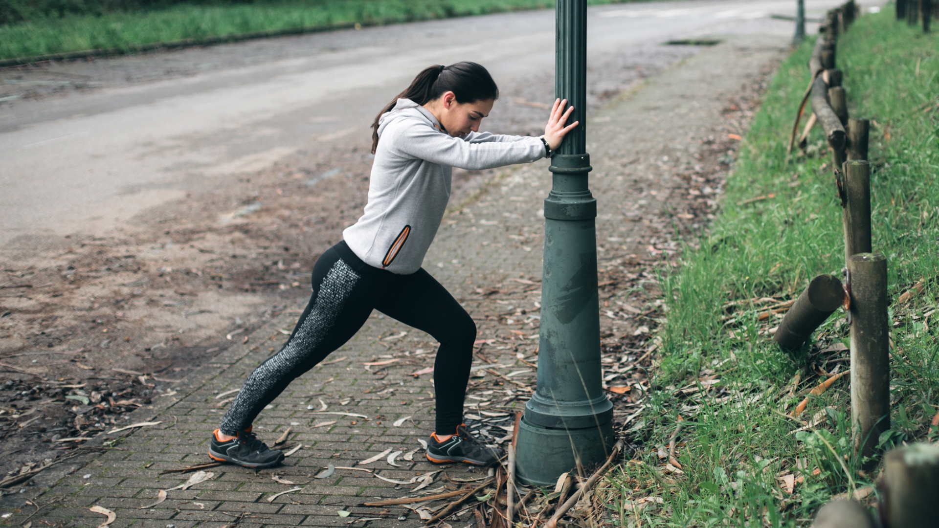 woman in leggings and hoodie doing a calf stretch against a lampost in a park setting