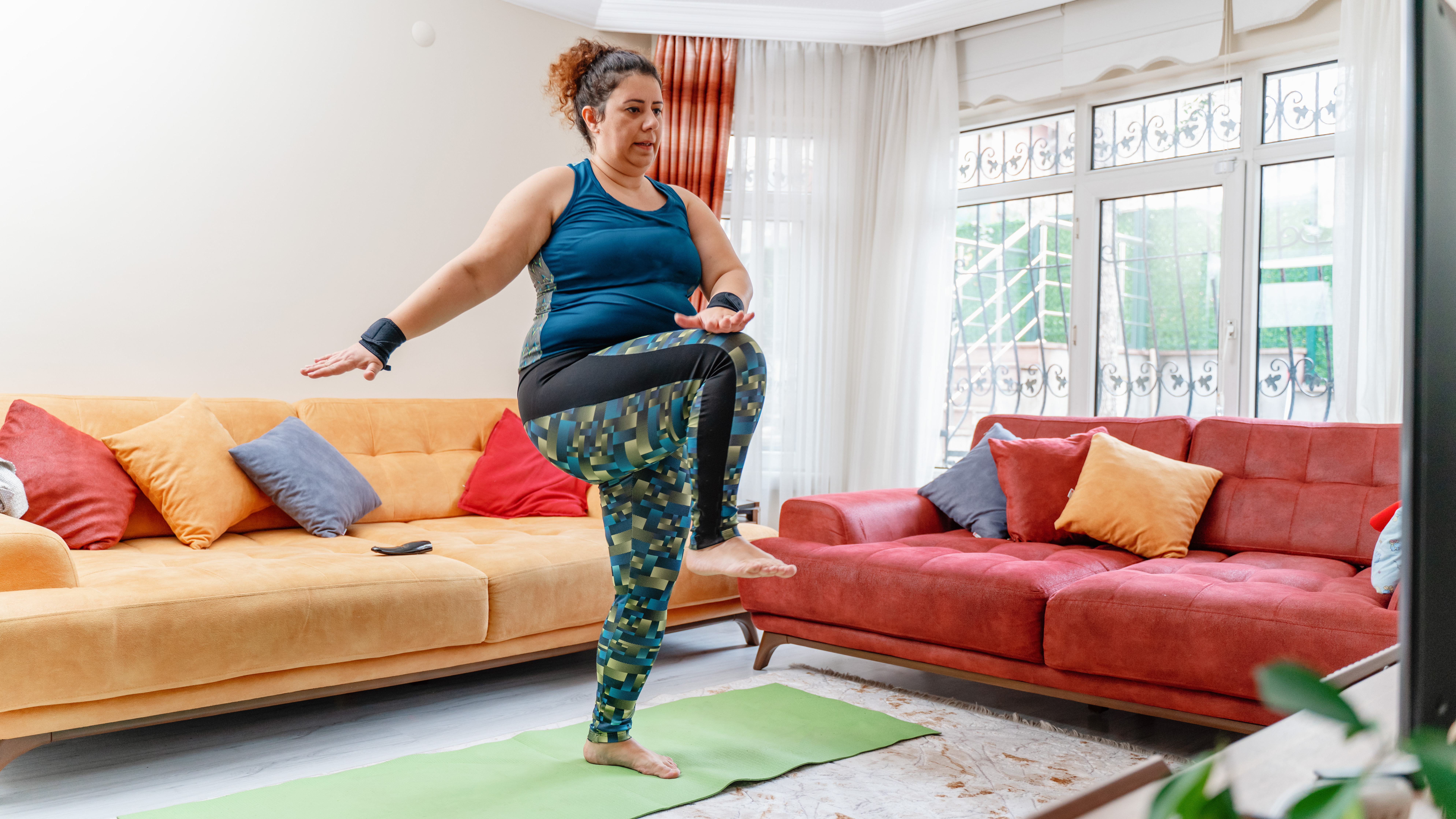 A woman works out at home in leggings and a vest. She is standing on an exercise mat, marching, with her right leg raised and her left hand touching her right knee. Behind we see two couches.