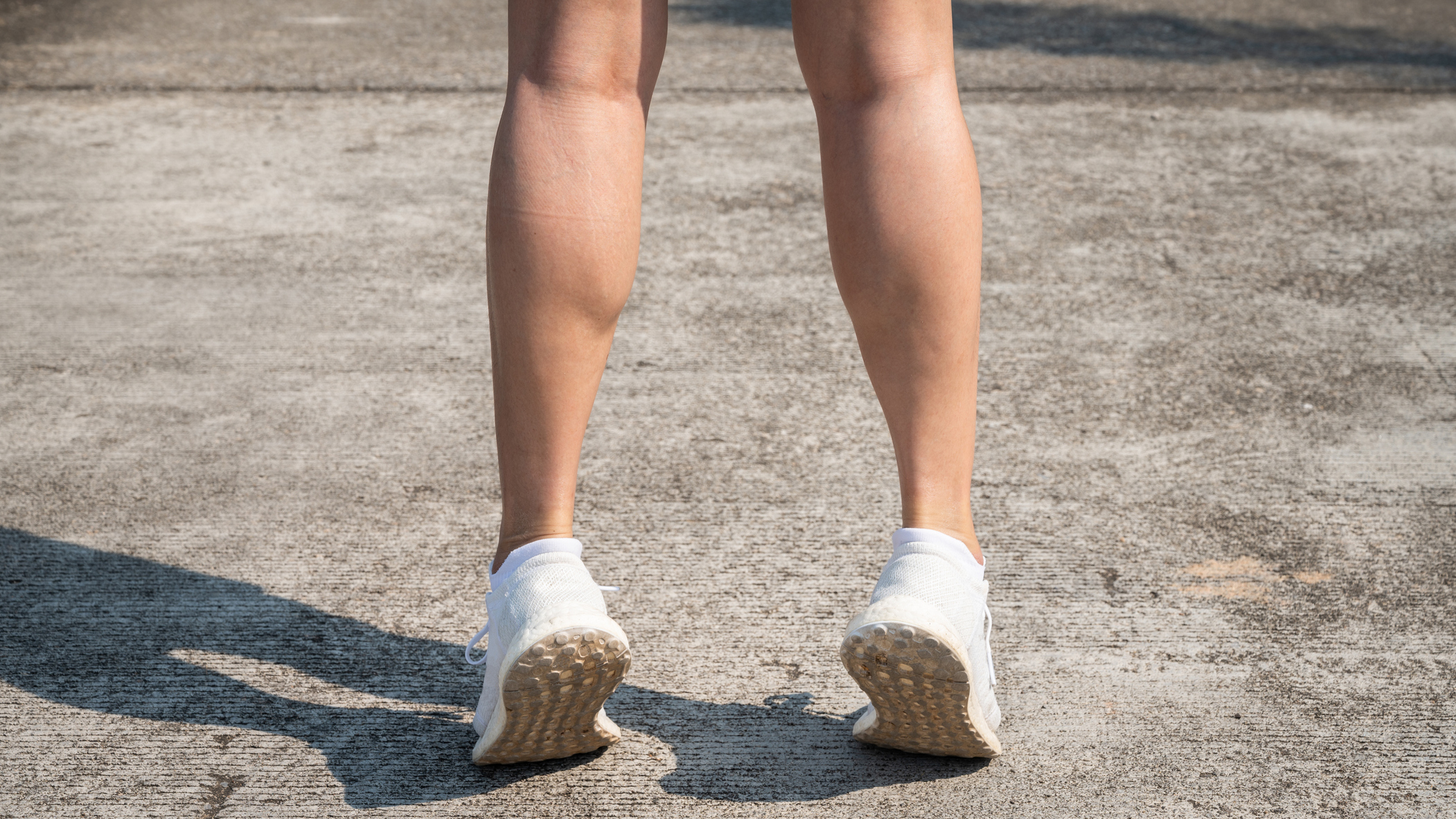 Person's bare legs below the knees seen from behind. They stand on tip toes and are wearing white trainers