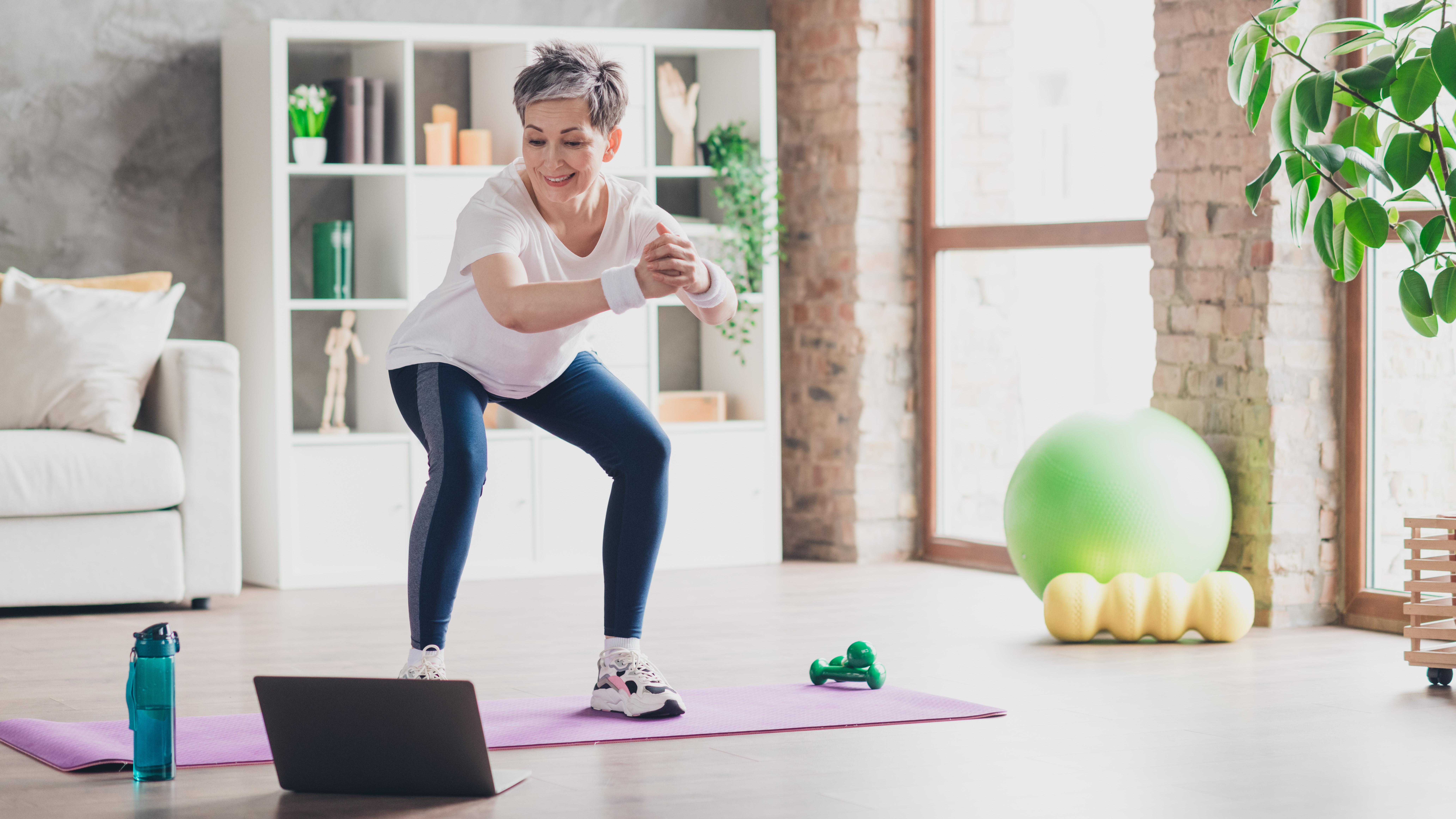 A woman performs a bodyweight squat at home on an exercise mat, while watching an open laptop at her feet. We see a water bottle, light dumbbells, an exercise ball and a foam roller in the room, along with a couch and bookshelves.