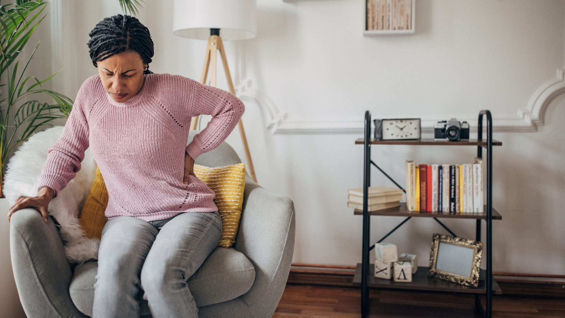 woman half seated on an armchair holding the side of her back looking pained. she's in a living room setting. 