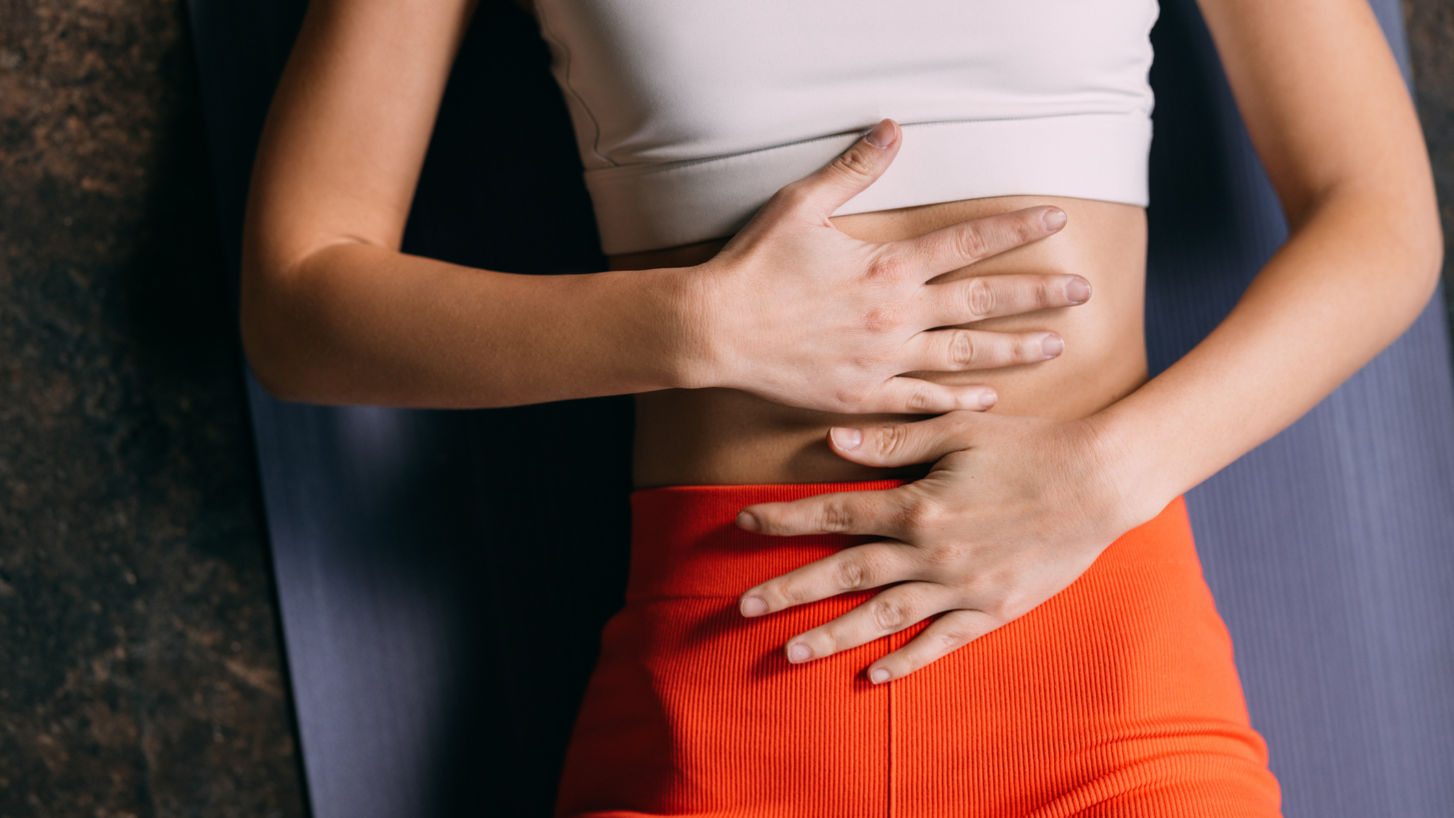 Woman in sportswear lies on her back on a yoga mat, with her hands over her midsection