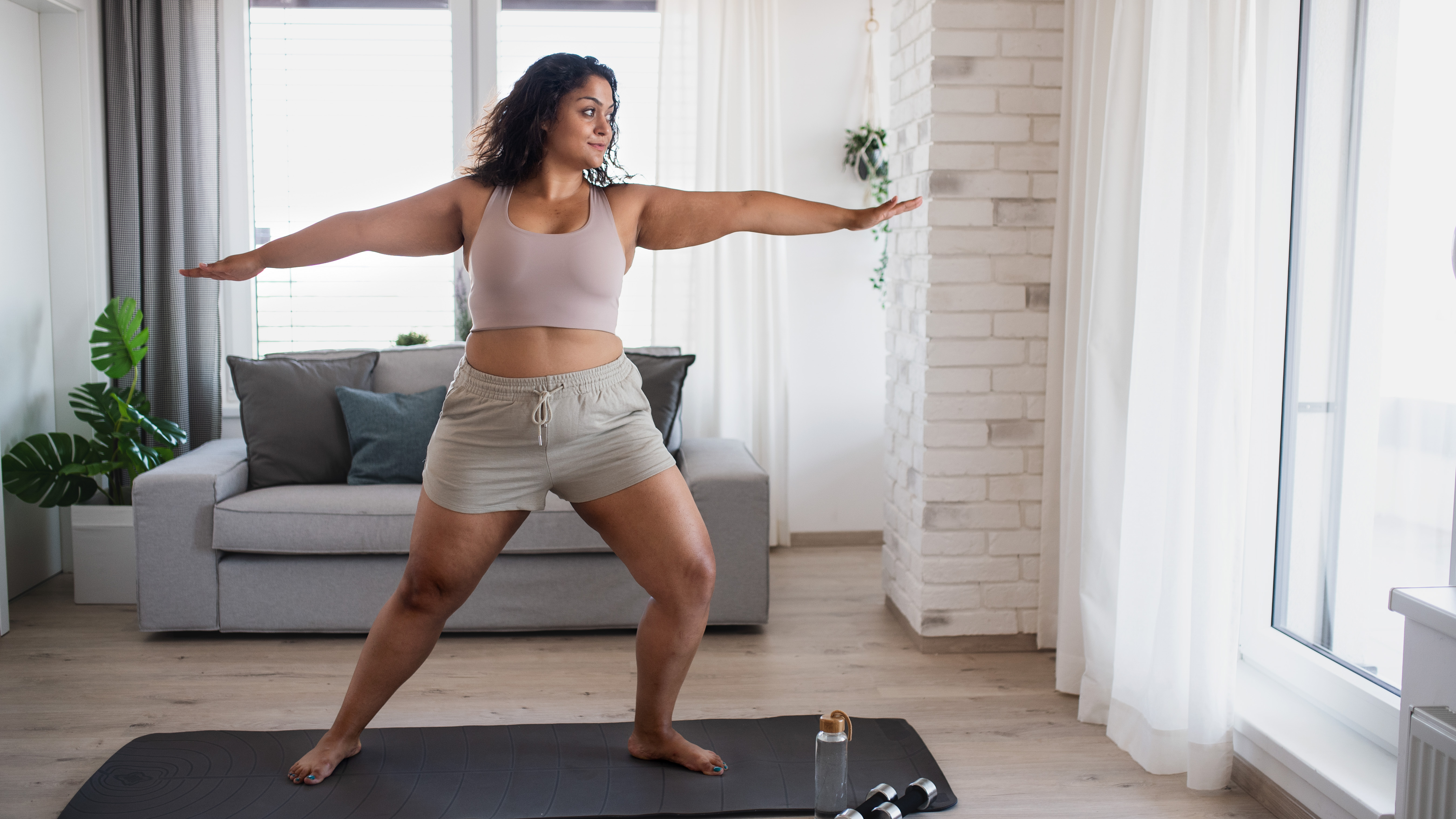 A woman performs a lunge in a yoga routine. She is in a living room, practicing on a yoga mat, with a couch and plant behind her. 