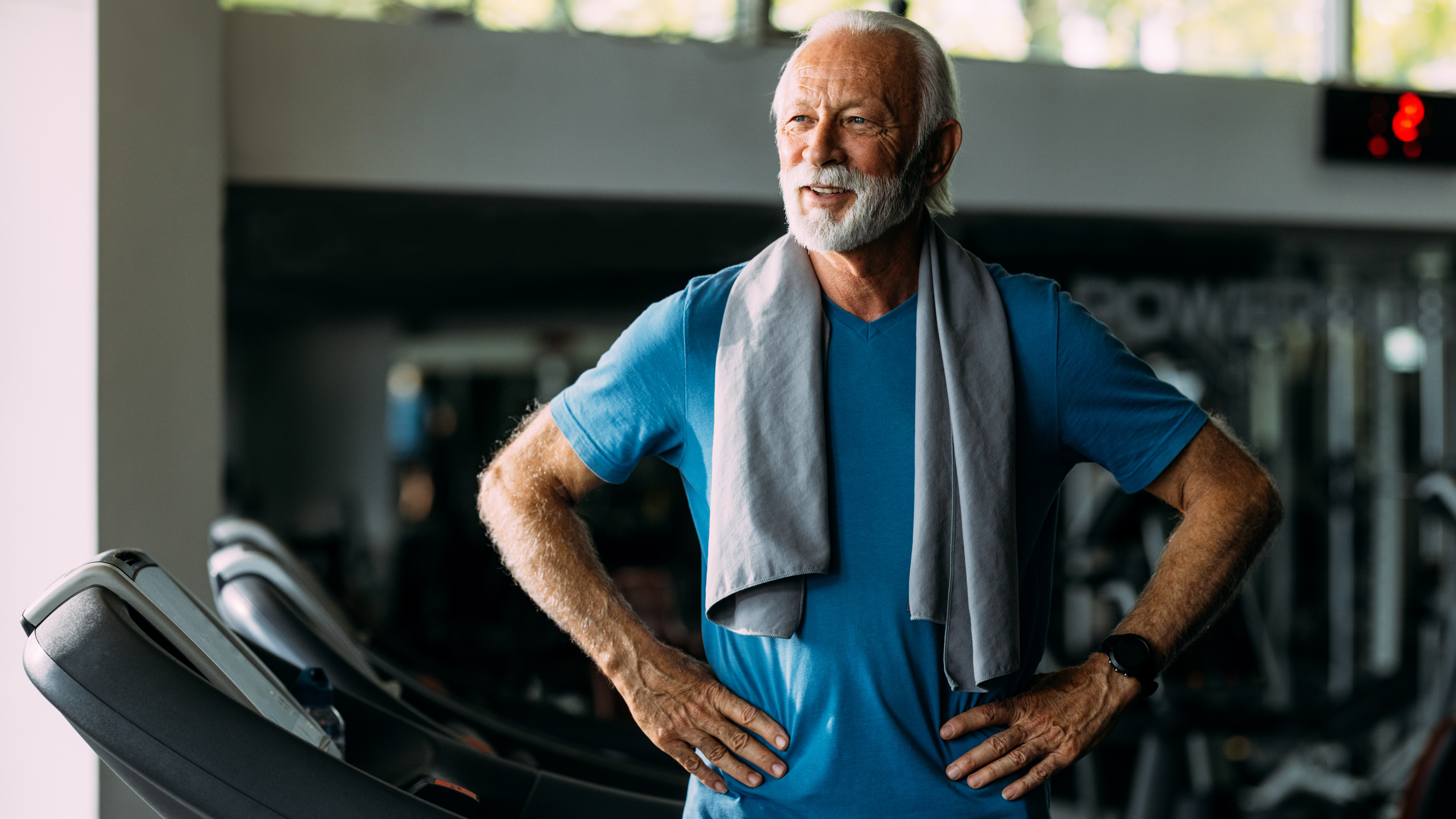 A man stands on a treadmill in a gym, with his hands on hips and a sweat towel draped over his shoulders. 